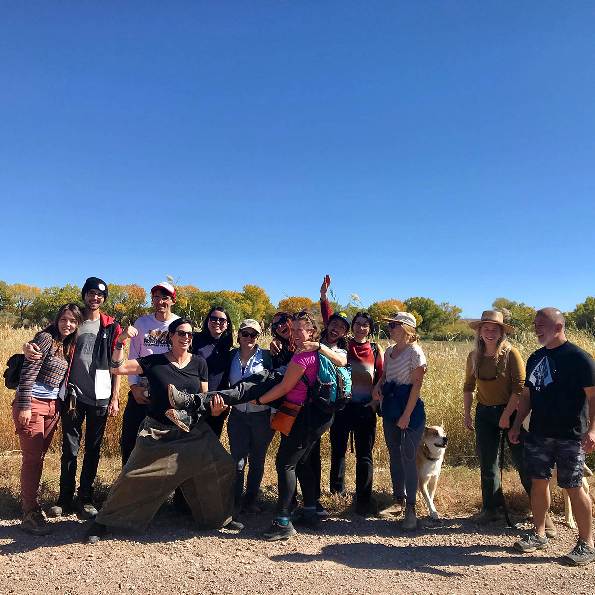 13 students pose in front of a grassy field with yellowing cottonwoods and clear blue sky behind them